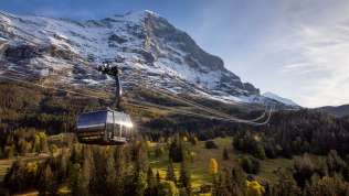 La funivia Eiger Express in primavera, con il paesaggio verdeggiante in primo piano, le montagne innevate sullo sfondo e il cielo azzurro.